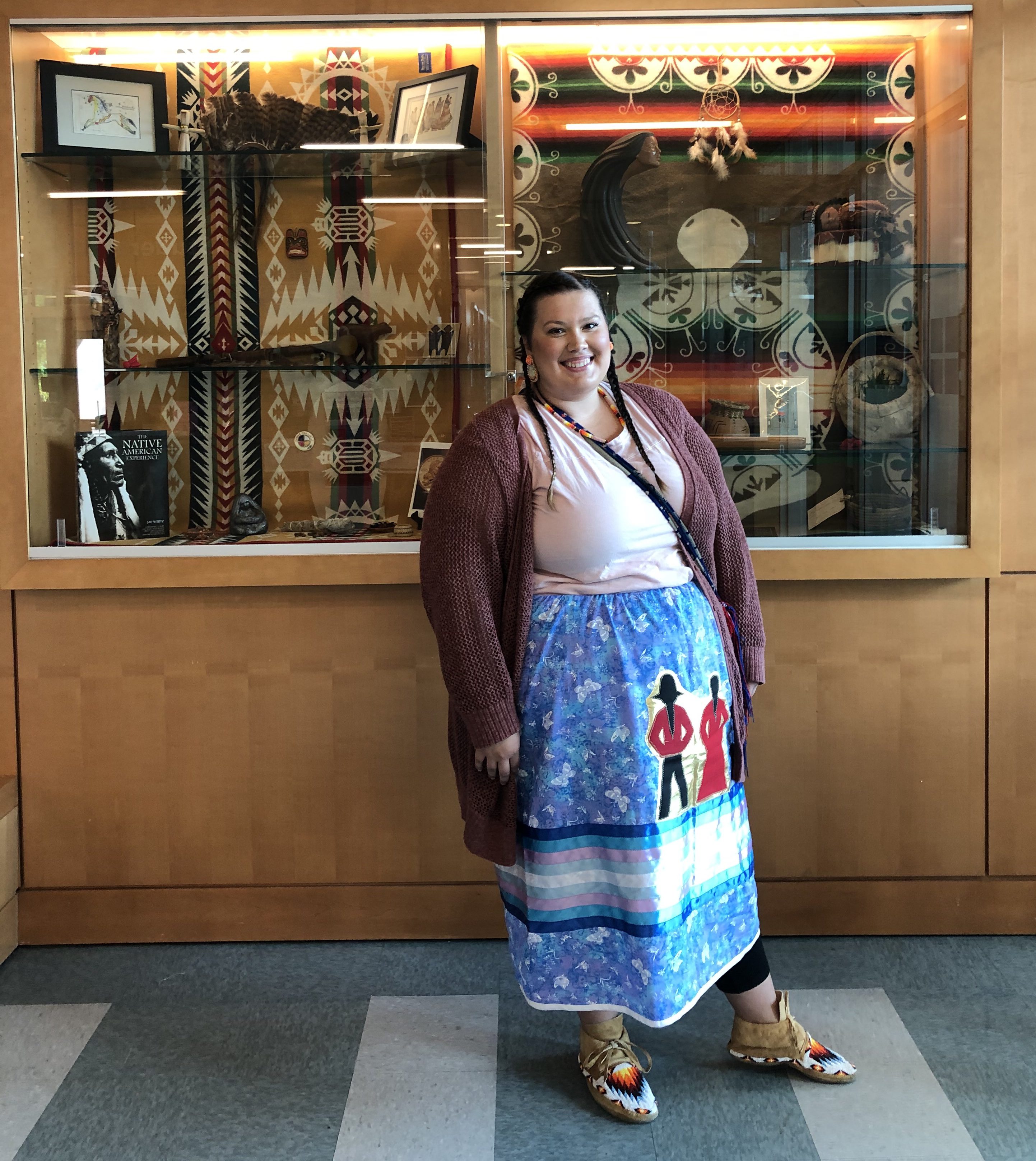 This shows a smiling Monique Lewis, Indigenous hairstylist, standing in front of a showcase filled with Indigenous artifacts.